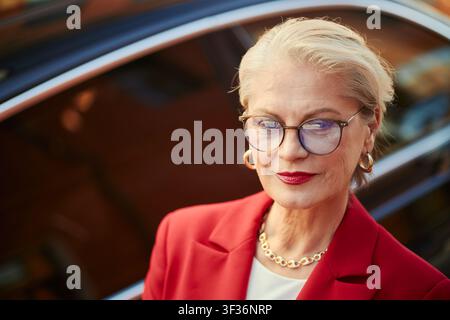 Portrait of senior Caucasian businesswoman standing by car, looking away and presenting executive confidence and mobility. Ideal for leadership, success, transport, luxury branding Stock Photo