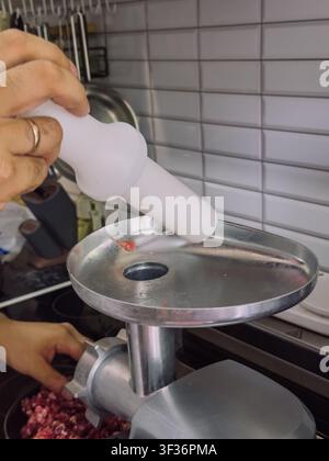 Using a meat grinder pusher to safely press raw meat into electric meat grinder while preparing homemade minced meat in home kitchen Stock Photo