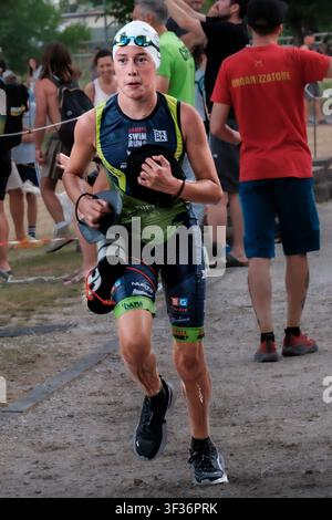 A focused young female competitor runs during the Ledro SwimRun Test Day at Lake Ledro, Italy, wearing a swim cap and holding hand paddles. Stock Photo