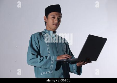 Muslim man in prayer attire holding a laptop and looking at the camera, isolated on a white background. Stock Photo