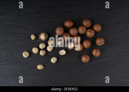 Macadamia nuts scattered. Natural raw peeled macadamia seeds and shells, the hardest and most expensive nuts in the world on black background top view Stock Photo