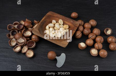 Macadamia nuts in wood bowl. Natural raw peeled macadamia seeds and shells, the hardest and most expensive nuts in the world on black background Stock Photo