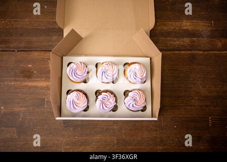 Box of six frosted cupcakes arranged in kraft bakery box on wooden table under warm light Stock Photo