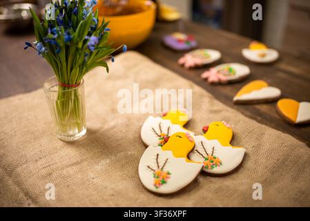 Handmade iced cookies shaped like chicks and eggs on rustic linen table with spring floral bouquet Stock Photo