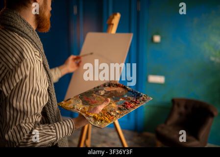 Artist at easel holding palette and brush in moody studio with textured blue walls and soft warm Stock Photo