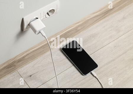 Smartphone charging from a white wall outlet with a power adapter and cable on a light wooden floor, the mobile device rests on a light wooden floor, Stock Photo