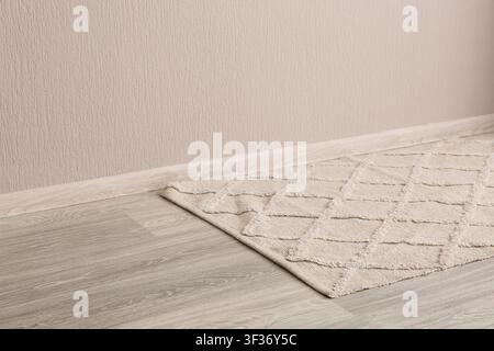 Beige textured rug with diamond pattern on light wooden floor against a neutral wall with baseboard, a neutral-colored wall with a matching baseboard Stock Photo