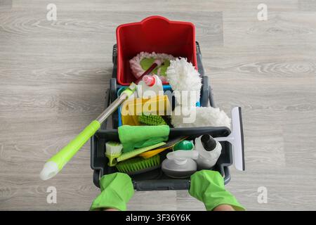 Person in green gloves pushing a cleaning cart filled with various household cleaning supplies and tools Stock Photo