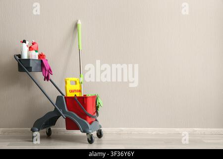 Professional cleaning cart with mop, bucket, and various cleaning supplies against a neutral wall Stock Photo
