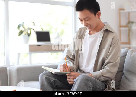 Young Asian man writing something in notebook on sofa at home Stock Photo