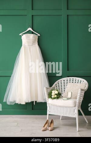 Beautiful wedding dress on green wall, wedding shoes on floor and bridal bouquet on white chair in room Stock Photo
