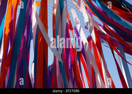 Low angle view looking up at vibrant, multicolored satin ribbons hanging from a wire mesh grid against a clear blue sky. Stock Photo