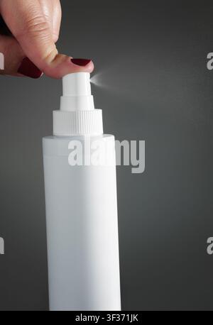 A woman finger pressing nozzle of a blank white plastic spray bottle spraying mist over dark background. Stock Photo