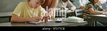 Header of Teen Boy Writing Notes with Teacher Support in School Classroom Stock Photo