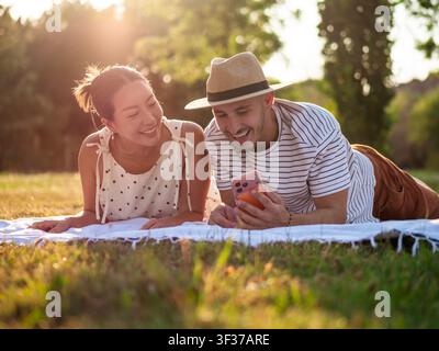 Happy multiracial couple is spending time together in park in summertime afternoon. They are using smartphone. Stock Photo