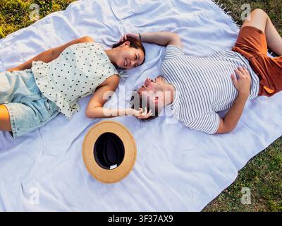 A happy couple lies on a picnic blanket in a peaceful park, smiling and talking as they enjoy a relaxing afternoon together. Stock Photo