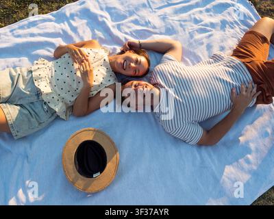An overhead view of a happy couple relaxing on a picnic blanket during golden hour, sharing smiles in a tranquil natural setting. Stock Photo