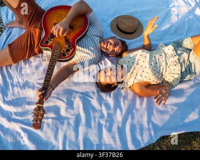 A smiling couple lies on a picnic blanket, playing guitar and singing. The warm golden light and carefree atmosphere capture the joy of music, friends Stock Photo