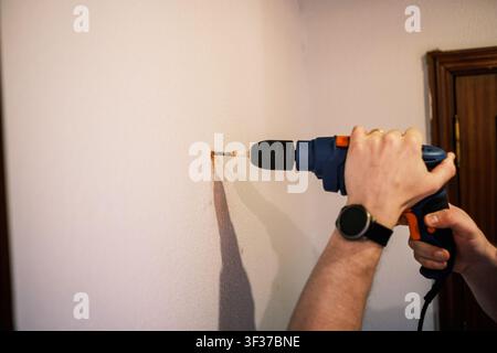 Person drilling a hole into a white interior wall using a blue power drill Stock Photo