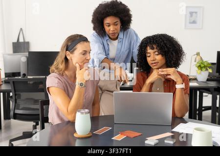 Diverse female coworkers collaborating around round table in bright office with laptop, swatches. Collaboration, team, designers, creative, workspace, monitors, samples Stock Photo