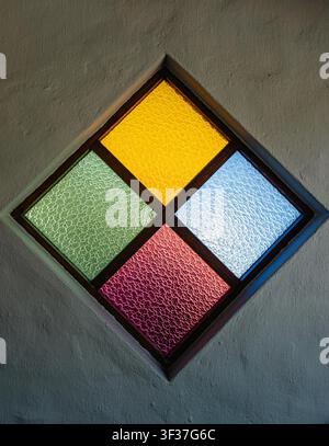 rhombus shape stained glass window is embedded in gray wall rough, textured surface. Window divided into four equal quadrats with textured, rippled glass different color, green, yellow, purple, blue Stock Photo