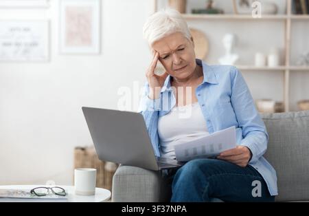 Thoughtful Elderly Woman Working With Papers And Laptop At Home, Checking Bills Stock Photo