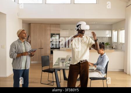 Diverse male coworkers testing VR headset, watching tablet, typing on laptop in kitchen Stock Photo