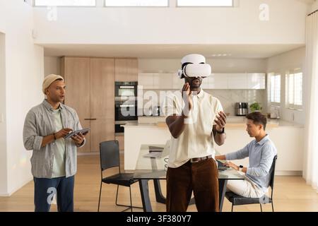 Diverse male coworkers testing white VR headset, using tablet and laptop in open-plan kitchen Stock Photo