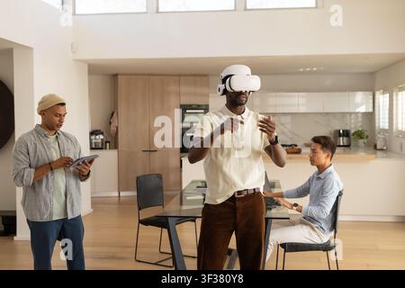 Diverse male coworkers testing white VR headset while using tablet and laptop in kitchen Stock Photo