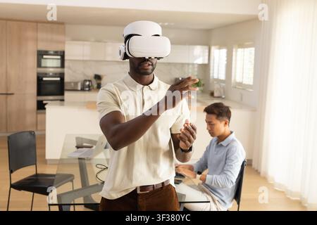 Diverse male colleagues testing VR wearing white headset and typing on laptop in kitchen Stock Photo