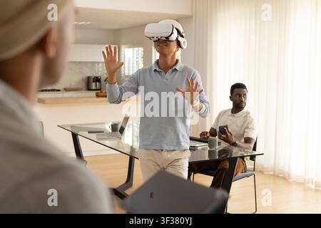 Diverse male team watching Asian man wearing white VR headset at glass table, holding smartphone Stock Photo