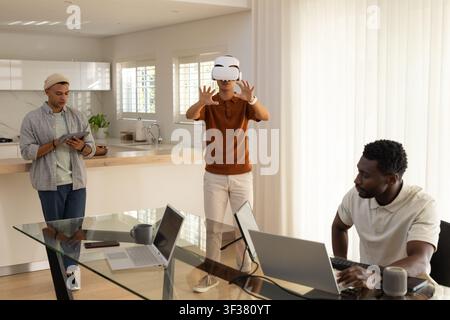 Diverse male coworkers in kitchen using laptops tablet Asian man wearing white VR headset reaching Stock Photo