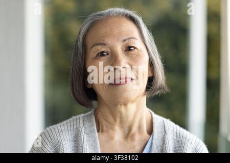 Senior Asian woman standing on porch wearing gray cardigan, blue top, stud earrings, garden behind Stock Photo