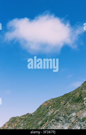 A white puffy cloud in a bright blue sky with a section of mountain in the foreground. Stock Photo