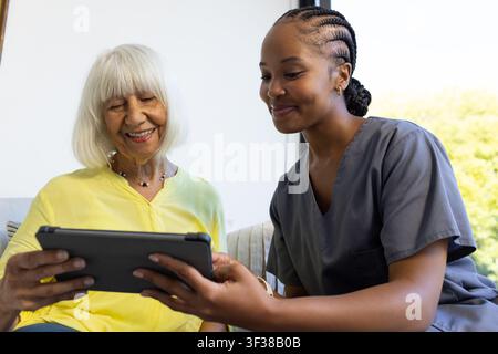 Senior woman in yellow blouse and female caregiver in scrubs sitting on sofa holding tablet Stock Photo