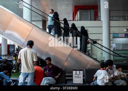 A group of people sit on a bench while others ascend an escalator in a modern indoor space. Stock Photo