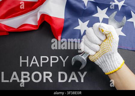 A gloved hand firmly holding a wrench against an American flag background with Happy Labor Day text on dark surface. High Quality Photo Stock Photo