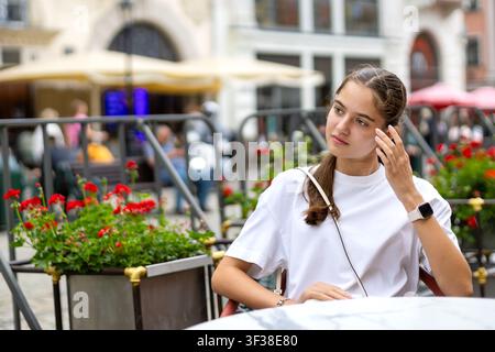 Young Woman Sitting at an Outdoor Cafe. Young woman in a white shirt and smartwatch sitting at an outdoor cafe, looking to the side with one hand Stock Photo