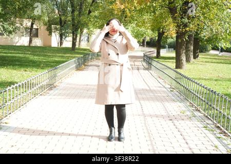 Woman adjusts coat on paved pathway. Trees line walkway behind her. Sunlight filters through green leaves. Metal railings flank both sides. Her boots Stock Photo