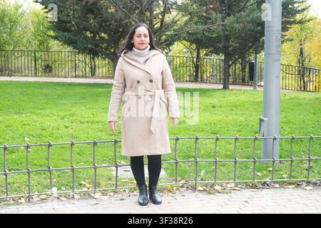 Woman stands in park wearing beige coat. She wears black leggings and boots. Green grass and trees surround her. Metal fence lies in foreground. Soft Stock Photo