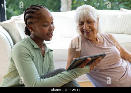 African American woman in green top holding tablet and senior woman smiling sitting in living room Stock Photo