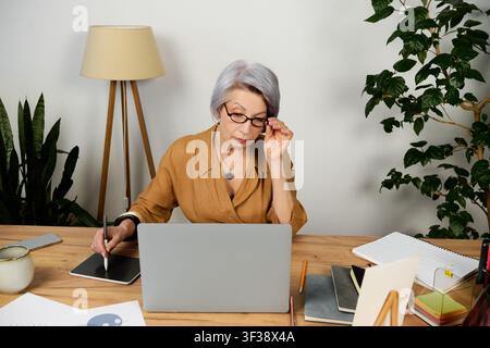 Senior businesswoman reviews documents while using a laptop and graphic tablet Stock Photo