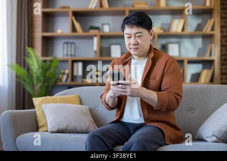 Asian man sitting on a sofa at home, relaxing while using his smartphone for texting, browsing, and connecting with social media Stock Photo