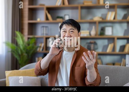 Smiling asian man having a cheerful phone conversation while relaxing on a sofa in a modern home living room or remote workspace, actively communicating with hand gestures Stock Photo