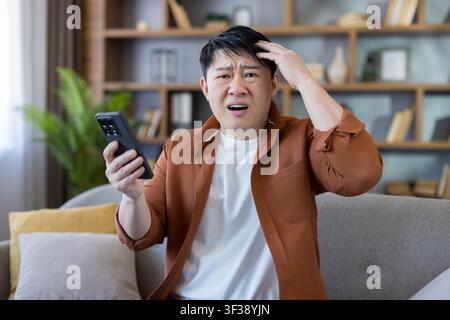Asian man looking confused and stressed, holding a smartphone while receiving shocking bad news or experiencing a digital mistake causing frustration Stock Photo