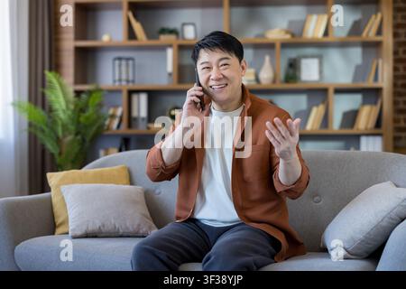 Asian man sitting on a sofa, happily talking on his mobile phone and gesturing with his hand in a modern living room, communicating and interacting remotely from home Stock Photo
