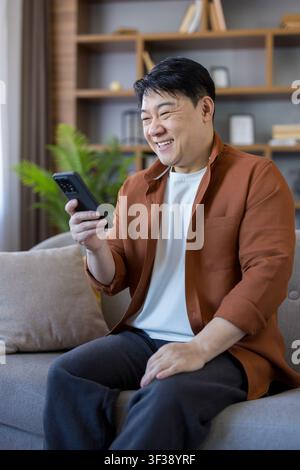 Cheerful asian man relaxing on a living room sofa, happily using his smartphone, connecting with friends and enjoying digital content during leisure time Stock Photo