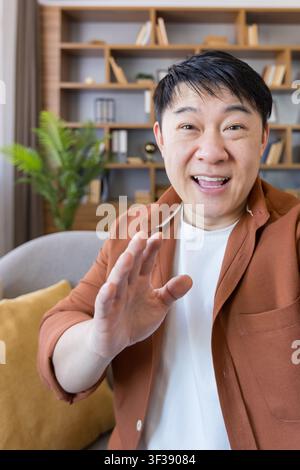 Cheerful asian man waving and greeting during a virtual video call from his home with a bookshelf in the background, communicating remotely Stock Photo