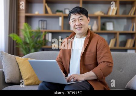 Asian man smiling, sitting comfortably on a sofa in his living room, looking at the camera while using a laptop for remote work or online studying from home Stock Photo