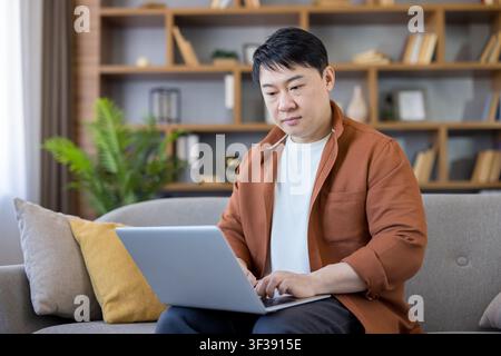 Asian man sitting comfortably on sofa, typing on laptop, engaging in remote work, online learning, or casual browsing at home Stock Photo